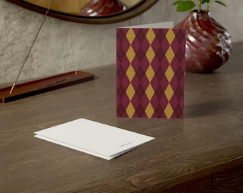 May include: A folded greeting card featuring a repeating argyle pattern in shades of deep burgundy and mustard yellow. A second, plain white card lies flat in front of the folded card, displaying a small, centered text logo on its surface.