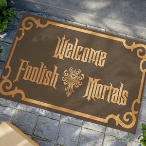 May include: A brown doormat with a gold border and the text "Welcome Foolish Mortals" in a gothic font. A gold ornate design is in the centre of the mat.