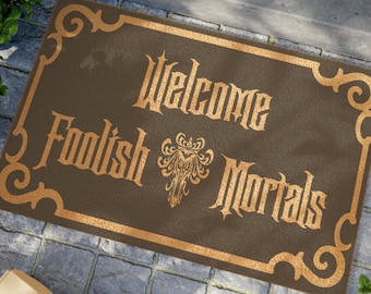 May include: A brown doormat with a gold border and the text "Welcome Foolish Mortals" in a gothic font. A gold ornate design is in the center of the mat.