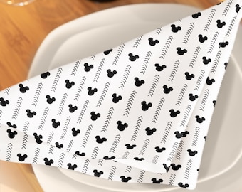 May include: White cloth napkin with a black Mickey Mouse head and arrow pattern, folded on a white plate. The napkin is part of a table setting, with a wine bottle and glass visible in the background.