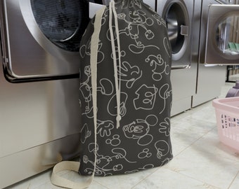 May include: A black laundry bag with a white drawstring and a pattern of Mickey Mouse outlines. The bag is sitting in front of a washing machine.