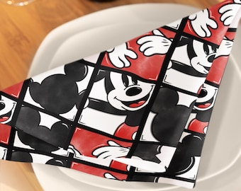 May include: A folded napkin with a repeating pattern of Mickey Mouse's face and hands. The design features black, white, and red squares. The napkin is placed on a white plate, with a wooden tray and wine bottle in the background.