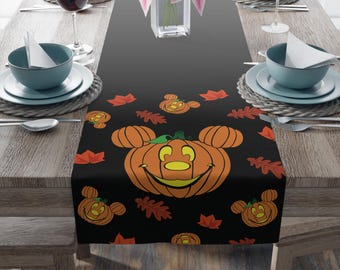 May include: A black table runner featuring a repeating pattern of orange jack-o'-lanterns with round ears and scattered red and orange autumn leaves. The design includes a large central pumpkin face with a wide smile and smaller pumpkin motifs.