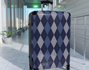 May include: A rolling suitcase with a blue, gray, and navy argyle pattern. The suitcase has a retractable handle and four wheels. The luggage is in a hallway with white tile flooring and potted plants.