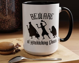 May include: A white ceramic mug with a black interior and handle. The front features a black silhouette graphic of three ghosts and the text "BEWARE of Hitchhiking Ghosts" in a stylized gothic font. The mug is cylindrical with a smooth finish.