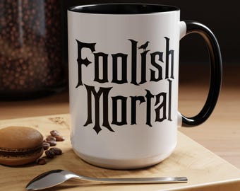 May include: A white ceramic mug with a black interior and handle, featuring the black text "Foolish Mortal" in a gothic-style font on the front. A small black bird silhouette is positioned above the word Foolish on the mug surface.