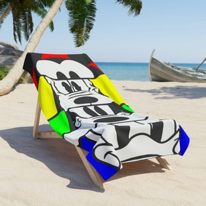 Puede incluir: Una toalla de playa con un diseño de personaje de dibujos animados, colocada sobre una silla de playa de madera. La toalla presenta un diseño de personaje en blanco y negro con acentos rojos, amarillos, verdes y azules. La escena está ambientada en una playa de arena con un barco al fondo.