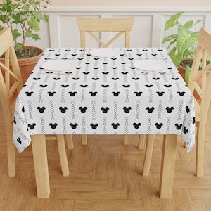 May include: A white tablecloth with a black and white pattern of Mickey Mouse heads and chevron lines. The tablecloth is laid on a wooden table with four wooden chairs.