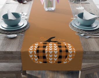 May include: A brown table runner featuring a pumpkin design. The pumpkin has a black stem and is decorated with a plaid pattern in orange and black, and white Mickey Mouse silhouettes. The runner is set on a wooden table with place settings.