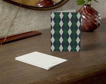 May include: A folded greeting card featuring a repeating argyle pattern in shades of dark green, charcoal gray, and white. A second, plain white greeting card lies flat in the foreground, showing a small, subtle logo printed on the bottom right corner.