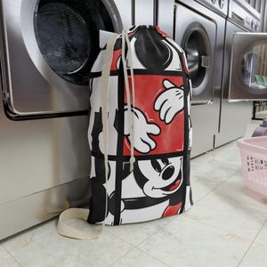 May include: A drawstring laundry bag featuring a repeating grid pattern of Mickey Mouse graphics in red, black, and white. The bag has a white fabric strap and a drawstring closure, displaying multiple close-up views of the character's face and white gloved hands.
