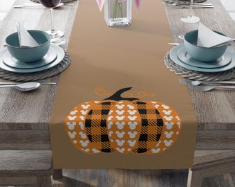 May include: A tan table runner featuring a central graphic of a pumpkin with alternating orange and black buffalo plaid sections and white mouse head silhouettes. The pumpkin has a black stem and orange swirling vine accents at the top.