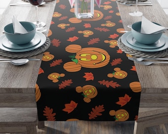 May include: A black table runner featuring a repeating pattern of orange pumpkins shaped like mouse ears with carved faces, interspersed with orange and red autumn leaves. The design includes various pumpkin sizes and leaf shapes on a dark background.