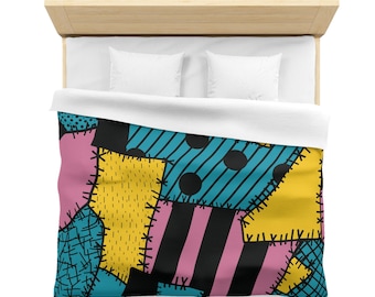 May include: A colorful patchwork duvet cover featuring a variety of geometric patterns. The design includes squares and rectangles in pink, yellow, teal, and black, with stitched details. The bed is made up with white pillows and a white sheet.
