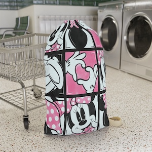 Puede incluir: Un cesto de ropa rosa y negro con una imagen de dibujos animados de Minnie Mouse. El cesto tiene un cierre de cordón y un asa blanca.