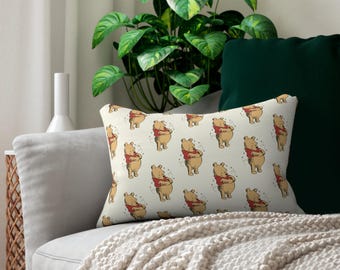 May include: Rectangular decorative pillow featuring a repeating pattern of a cartoon bear wearing a red shirt. The bear is standing with its arms crossed. The pillow is on a gray sofa with a white blanket and green pillows.