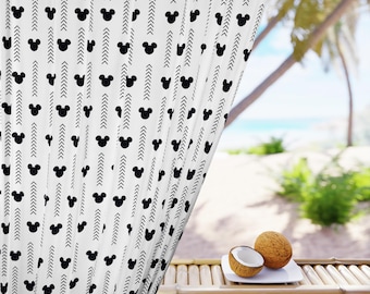 May include: White curtain with a black Mickey Mouse head pattern and arrow design. The curtain is hanging in front of a tropical beach scene. Two coconuts are on a white plate on a bamboo table.