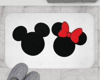May include: A rectangular white bath mat featuring two black mouse-ear silhouettes. The silhouette on the left is plain, while the silhouette on the right is adorned with a bright red bow between the ears. The mat has rounded corners.