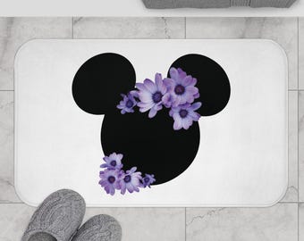 May include: A rectangular white bath mat featuring a large black silhouette of a mouse head shape. The silhouette is decorated with clusters of light purple daisy-like flowers positioned along the top and bottom left edges of the black shape.