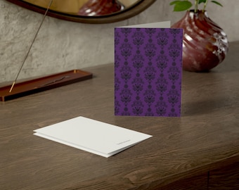 May include: A folded greeting card featuring a vibrant purple background with a repeating black damask pattern. A second, plain white greeting card lies flat in the foreground, showing its blank surface and a small, subtle logo on the bottom edge.