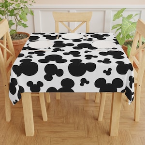 May include: A white tablecloth with a black Mickey Mouse pattern. The tablecloth is on a wooden table with four chairs.