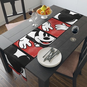 May include: A black and white table runner with a red background featuring a cartoon image of Mickey Mouse. The runner is on a black wooden table with a white plate and silverware.