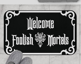 May include: A rectangular black doormat with a white decorative border and the white text Welcome Foolish Mortals. Between the words Foolish and Mortals is a white ornate graphic design. The mat has rounded corners and a flat surface texture.