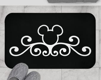 May include: A rectangular black bath mat featuring a centered white graphic design. The design consists of a stylized mouse head silhouette positioned above symmetrical, intricate white scrollwork patterns that extend horizontally across the mat.