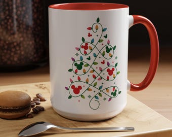 May include: A white ceramic mug with a red interior and handle, featuring a graphic of a Christmas tree formed by green swirling lines, colorful holiday light bulbs, and red and green Mickey Mouse head silhouettes on the front surface.