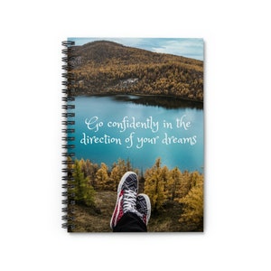 May include: Spiral-bound notebook featuring a scenic landscape with a lake and mountains. The cover displays the text "Go confidently in the direction of your dreams." The image includes a pair of sneakers with a black, white, and red design.