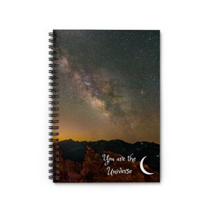 May include: Spiral-bound notebook featuring a night sky image with the Milky Way and mountains. The cover includes the text "You are the Universe" and a crescent moon graphic. The notebook has a black spiral binding.