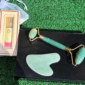 Puede incluir: Un conjunto de rodillo y gua sha de jade verde sobre un fondo negro. El rodillo tiene un mango dorado y el gua sha tiene forma de corazón. El conjunto está empaquetado en una caja negra con un borde dorado. La caja también incluye una pequeña botella de perfume etiquetada como "Root Roll On Perfume".