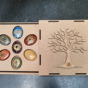 May include: A wooden box with a tree design on the lid, containing seven chakra stones with gold symbols. The stones are arranged in a circle and are in the colors of the chakras: red, orange, yellow, green, blue, indigo, and violet.