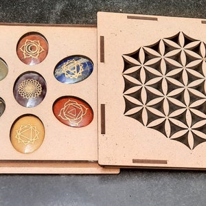 Op de afbeelding: Een houten doos met een levensbloemontwerp op de deksel. De doos bevat zeven chakrastenen met gouden symbolen.