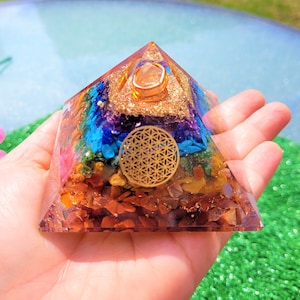 Pode incluir: Uma pirâmide transparente de resina com um design dourado de flor da vida no centro. A pirâmide está cheia de pequenos cristais coloridos, incluindo vermelho, laranja, amarelo, verde, azul, índigo e violeta. O topo da pirâmide é coroado com um cristal transparente.