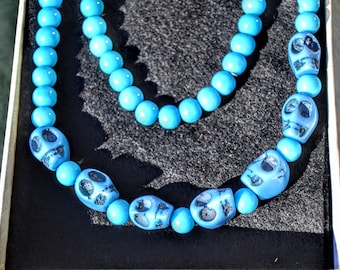 May include: A turquoise blue beaded necklace with skull charms. The necklace is displayed on a black surface.