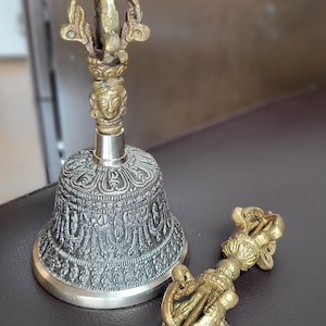 May include: A silver bell with intricate designs and a gold-colored handle with a stylized figure at the top. A matching gold-colored hand-held bell with a similar design is lying on its side.