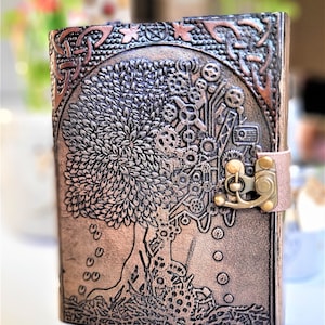 May include: A copper-toned leather journal with a detailed embossed design of a tree with gears and a Celtic knot border. The journal has a brass clasp and a leather strap.