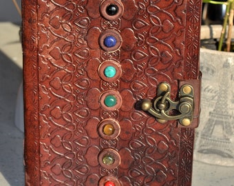 Puede incluir: Diario de cuero marrón con un diseño estampado decorativo y un cierre de metal. El diario tiene siete piedras preciosas de colores incrustadas en la portada.