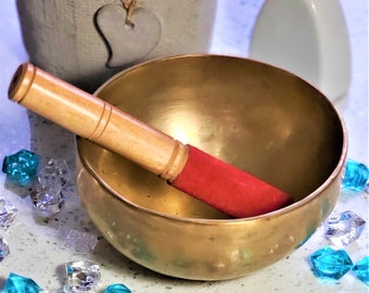 Puede incluir: Un cuenco cantor dorado con un mazo de madera con una punta de fieltro roja. El cuenco está sobre una superficie blanca con cristales azules y verdes.