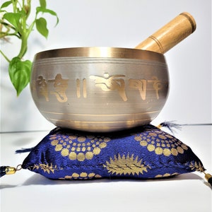 May include: A silver singing bowl with gold trim and etched symbols, on a blue cushion with a gold floral pattern. A wooden mallet rests inside the bowl. The cushion has tassels on each corner.