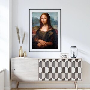 May include: A framed print of the Mona Lisa painting, featuring a woman with a soft smile and a dark background. The print is displayed on a light wood cabinet with a geometric pattern.