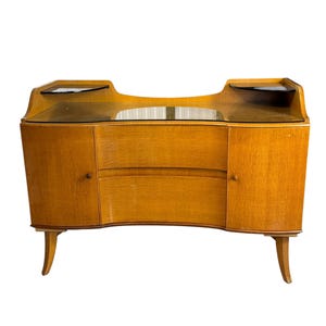 May include: A light brown wooden vanity with a curved front, featuring two side cabinets with small round knobs, a central drawer, and a dark glass top surface. The piece is supported by four tapered legs and includes two small elevated corner shelves.