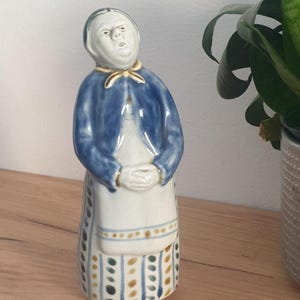 May include: A ceramic figurine of a person wearing a blue jacket, a white apron, and a long skirt with vertical blue and yellow dotted stripes. The figure has a gold-colored collar detail, hands clasped in front, and a neutral facial expression.