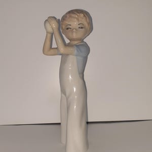 Può includere: Statuina in porcellana di un giovane in tuta bianca e top azzurro chiaro, che tiene una palla bianca. La statuina ha un'espressione neutra ed è in piedi. La figura è alta circa 20 cm.