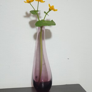 May include: A slender, pink glass vase with a dark purple base holds several yellow flowers with green stems and leaves. The vase is on a dark brown surface against a white wall.