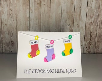 May include: A white card with the text "The stockings were hung" and three paper stockings hanging from a string. The stockings are red, purple, and yellow with the names "Mickey", "Minnie", and "Pluto" written on them.