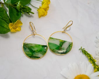 May include: A pair of gold hoop earrings with a half-circle design filled with green and white resin. The earrings have a hook closure and are adorned with small crystals. The image is styled with yellow flowers and green foliage.
