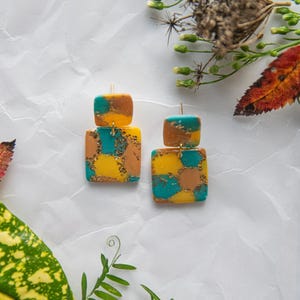 Op de afbeelding: Een paar hangende oorbellen met een vierkante vorm. De oorbellen zijn gemaakt van polymer clay en hebben een turquoise, gele en bruine kleurstelling met gouden glitters.