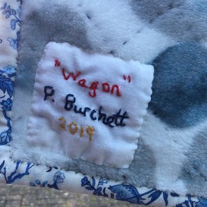 May include: A white fabric label with blue, red, and gold embroidery. The label reads "Wagon" P. Burchett 2019.